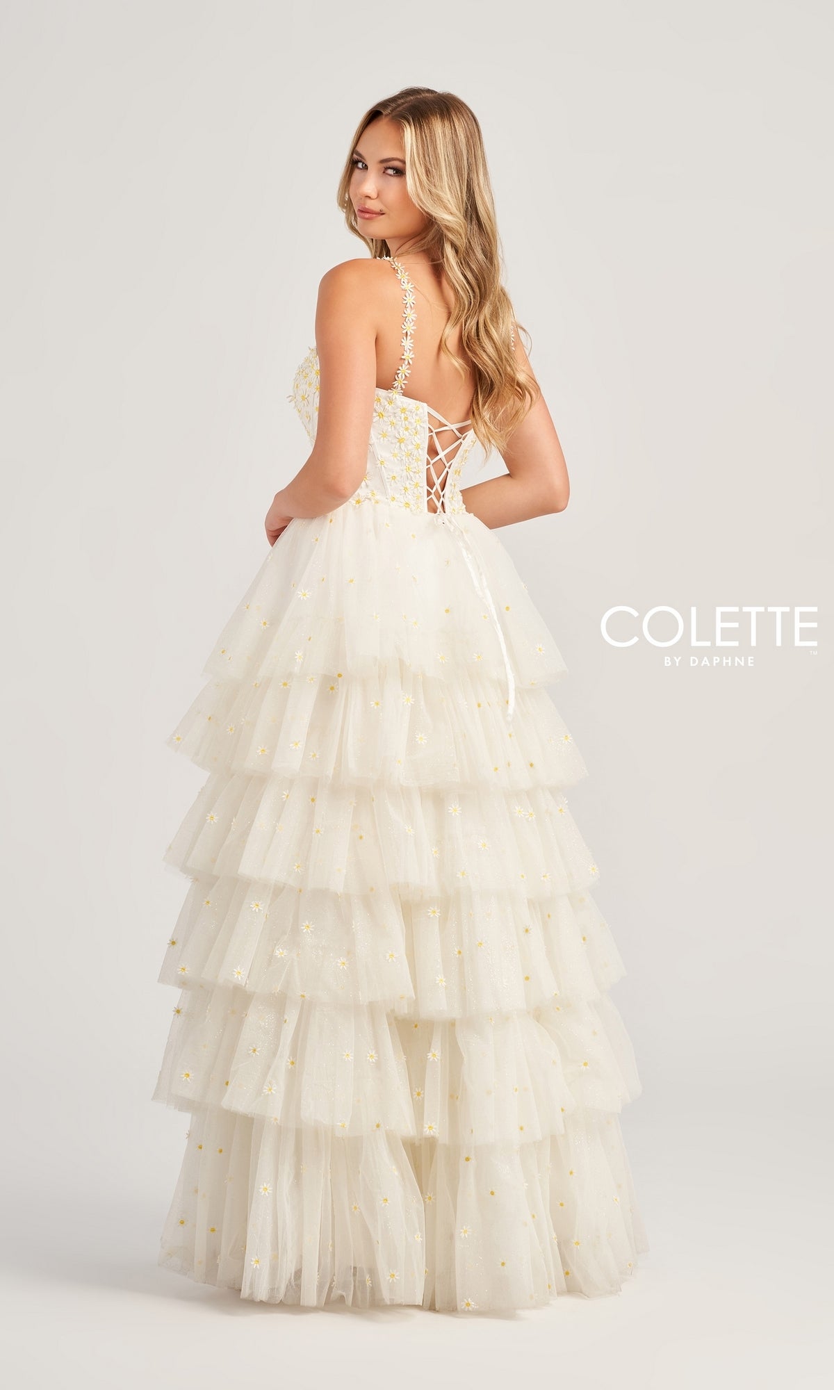 A woman wears the Colette High-Low Daisy Tiered Prom Dress CL5237, a white tulle gown with gold accents, lace-up back, and spaghetti straps, as she faces away and looks over her shoulder against a plain light gray background.